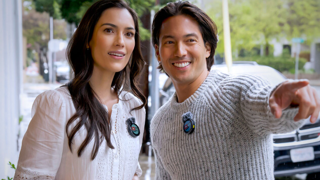 A smiling couple wearing wireless microphones, interacting in front of a car.