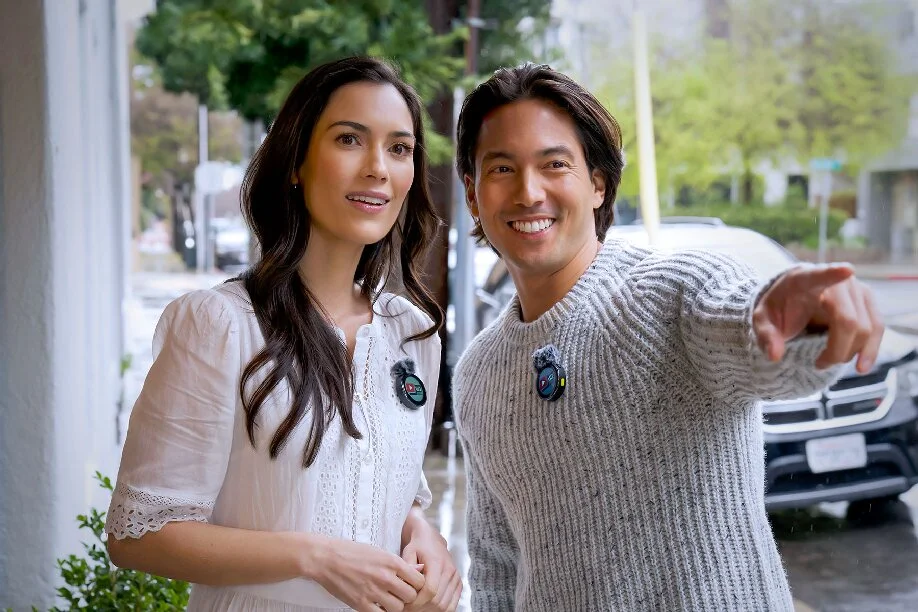 A man and woman, each wearing a clip-on lavalier microphone, stand outdoors while the man points ahead.