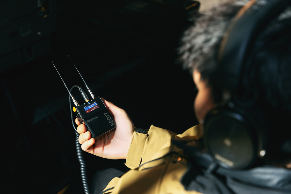 A person holds a Saramonic wireless audio transmitter, likely connected to a microphone, while wearing headphones.