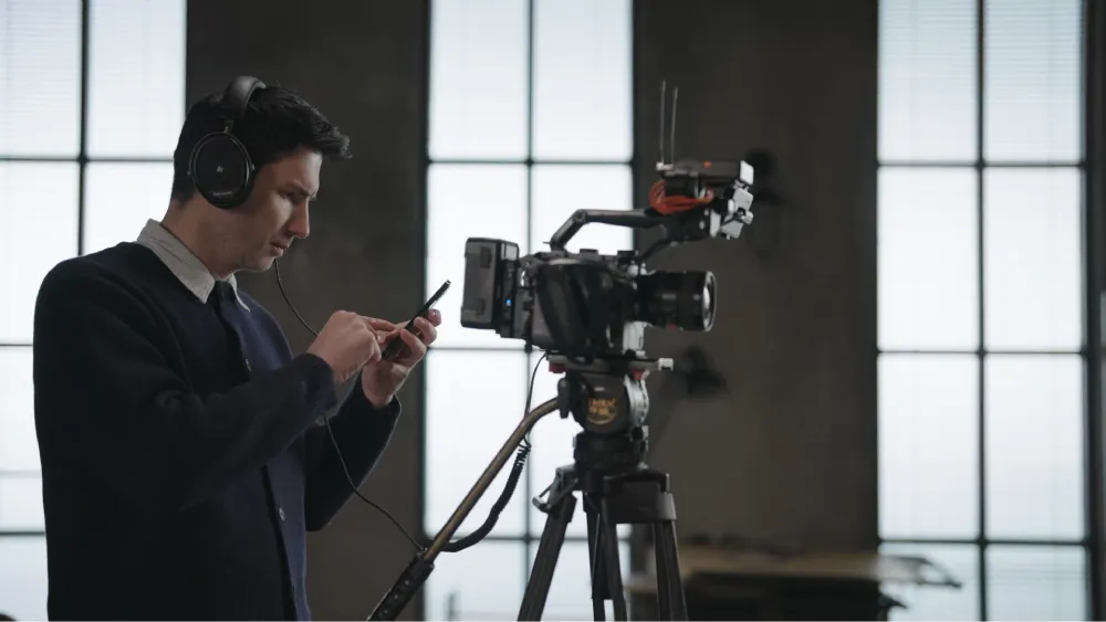 A sound engineer in headphones adjusts settings on a smartphone connected to a professional video camera equipped with a microphone, ready for filming.
