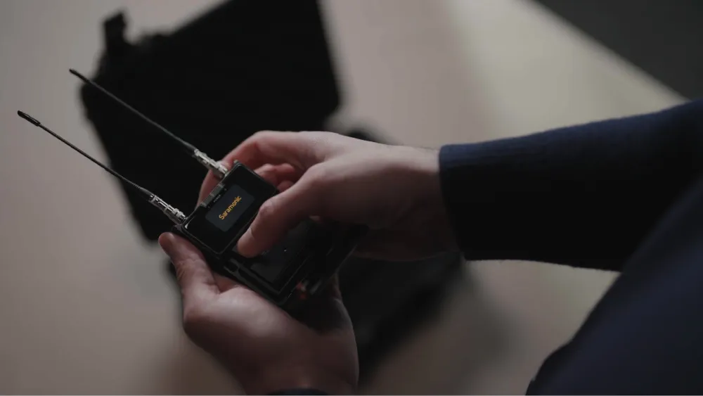 A person holds a Saramonic wireless microphone receiver, interacting with its screen and buttons, with an open case blurred in the background.