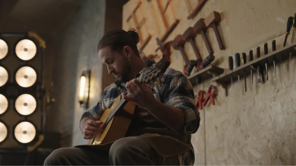 A man with a bun plays an acoustic guitar in a workshop with tools on the wall;  a recording or performance setup.