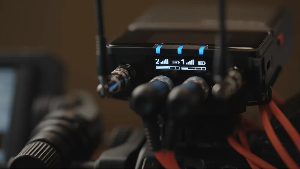 Close-up of a wireless microphone receiver displaying signal strength and battery levels for two audio channels, with connected cables and antennas visible.