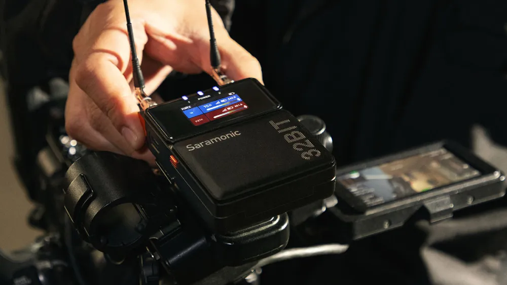 A hand adjusts a Saramonic wireless microphone receiver mounted on equipment, its screen illuminated, next to a phone serving as a monitor.