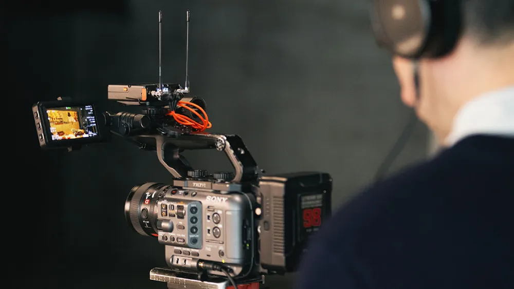 A cameraman, seen from behind and wearing headphones, operates a professional Sony video camera equipped with an external monitor and a wireless microphone receiver.