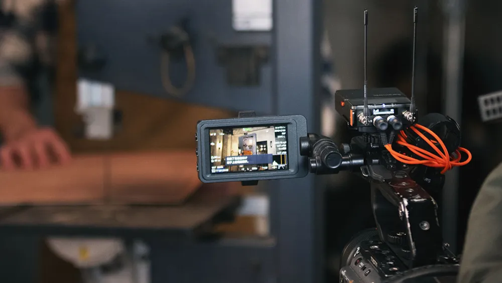 A video camera with an attached monitor and wireless microphone receiver is filming a person working with wood on a band saw in a workshop.